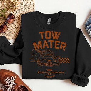 May include: Black sweatshirt featuring the text "TOW MATER" in orange, along with an illustration of a tow truck. The design includes the text "Piston Cup Racing Series" and "5 Time Champ". The sweatshirt is paired with blue jeans and brown shoes.