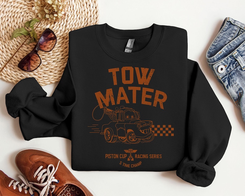 May include: Black sweatshirt featuring the text "TOW MATER" in orange, along with an illustration of a tow truck. The design includes the text "Piston Cup Racing Series" and "5 Time Champ". The sweatshirt is paired with blue jeans and brown shoes.