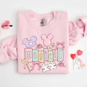 May include: A light pink sweatshirt with the word "magical" in pastel colors, featuring Mickey Mouse-inspired designs, bows, and a magic wand. Heart-shaped sunglasses and lollipops are also visible.