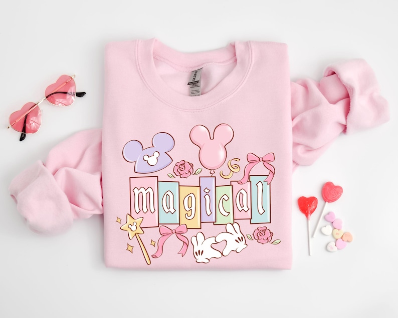 May include: A light pink sweatshirt with the word "magical" in pastel colors, featuring Mickey Mouse-inspired designs, bows, and a magic wand. Heart-shaped sunglasses and lollipops are also visible.
