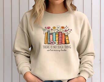 Puede incluir: Una sudadera beige con un gráfico de una pila de libros coloridos con flores en la parte superior. El texto "THERE IS NO SUCH THING as too many books" está impreso debajo del gráfico.