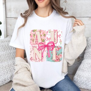 May include: White t-shirt featuring the word "AUNT" in pink letters with a pink bow. The letters are set against floral and patterned backgrounds in pink and teal. The shirt is a casual, everyday wear item.