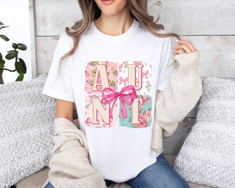 May include: White t-shirt featuring the word "AUNT" in pink letters with a pink bow. The letters are set against floral and patterned backgrounds in pink and teal. The shirt is a casual, everyday wear item.