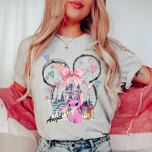 May include: A light gray t-shirt featuring a cartoon design of a pink character, a castle, and a pink bow. The design is framed by a Mickey Mouse head outline. The word "Angel" is written in script below the character.