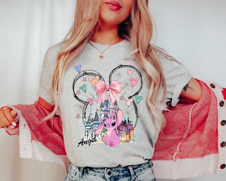 May include: A light gray t-shirt featuring a cartoon design of a pink character, a castle, and a pink bow. The design is framed by a Mickey Mouse head outline. The word "Angel" is written in script below the character.