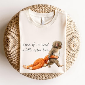 May include: Cream-colored t-shirt featuring a whimsical illustration of a brown monkey carrying an orange stuffed animal. The text "some of us need a little extra love" is printed in black script. The shirt is displayed on a woven, circular surface.