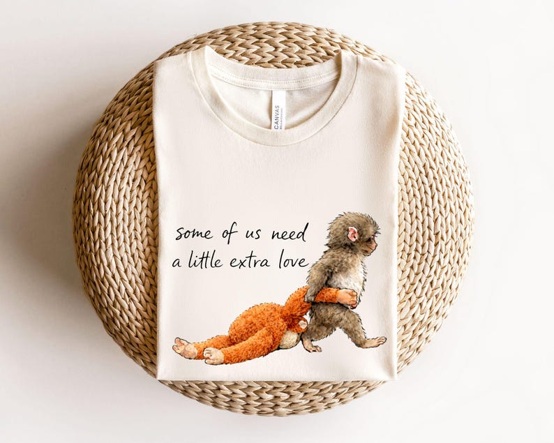 Peut inclure: T-shirt cr&egrave;me avec une illustration fantaisiste d'un singe brun portant un animal en peluche orange. Le texte "some of us need a little extra love" est imprim&eacute; en script noir. Le t-shirt est pr&eacute;sent&eacute; sur une surface circulaire tiss&eacute;e.