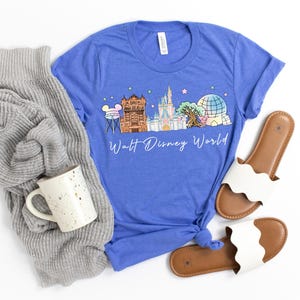 May include: A blue t-shirt featuring a colorful illustration of Walt Disney World landmarks, including Cinderella's castle and Spaceship Earth. The text "Walt Disney World" is written in a script font. The shirt is paired with a gray sweater, a speckled mug, and white sandals.