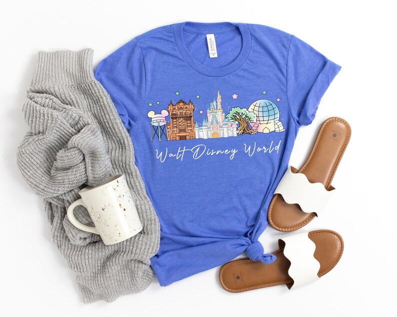 May include: A blue t-shirt featuring a colorful illustration of Walt Disney World landmarks, including Cinderella's castle and Spaceship Earth. The text "Walt Disney World" is written in a script font. The shirt is paired with a gray sweater, a speckled mug, and white sandals.