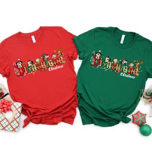 May include: Two short-sleeved t-shirts, one red and one green, featuring a festive "Disneyland Christmas" design. The design includes cartoon characters and holiday lights. The shirts are displayed on a white background with Christmas decorations.