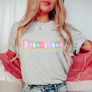 Puede incluir: Camiseta gris claro con la palabra "Disneyland" en colores pastel. El texto est&aacute; en un dise&ntilde;o de bloque rectangular con un borde dorado. La persona lleva un c&aacute;rdigan rojo y blanco.