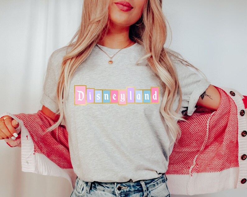 Puede incluir: Camiseta gris claro con la palabra "Disneyland" en colores pastel. El texto est&aacute; en un dise&ntilde;o de bloque rectangular con un borde dorado. La persona lleva un c&aacute;rdigan rojo y blanco.