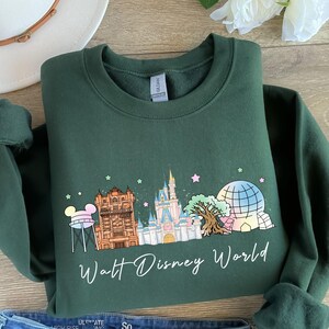 May include: Dark green sweatshirt featuring a colorful graphic of Walt Disney World landmarks, including Cinderella's castle and Spaceship Earth. The words "Walt Disney World" are written in a script font below the graphic.