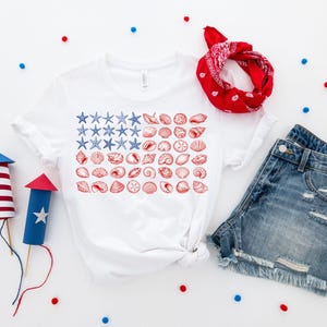 May include: A white short-sleeve t-shirt featuring a graphic of the American flag composed of blue starfish and red seashells. The set includes a red paisley bandana, blue denim shorts with frayed hems, and two decorative paper firework tubes.