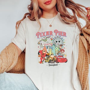 Puede incluir: Camiseta blanca con un gráfico colorido de personajes de Pixar en Pixar Pier en Disneyland. El gráfico incluye personajes como Buzz Lightyear, Woody, Mike Wazowski, Sulley, y más.