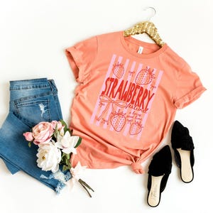 May include: A peach-coloured t-shirt with a strawberry graphic and the text "Strawberry Festival a berry good time." The shirt is paired with blue jeans, black shoes, and a bouquet of flowers. The shirt is hanging on a gold hanger.