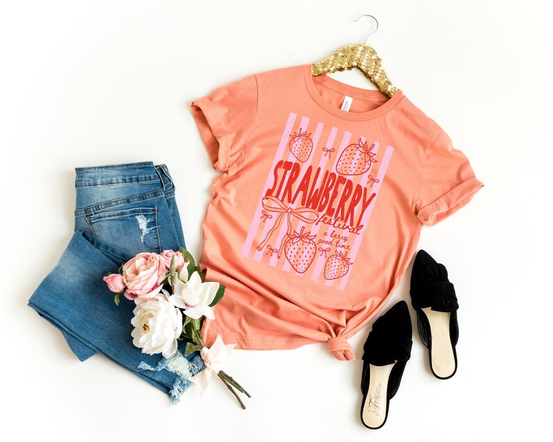 May include: A peach-coloured t-shirt with a strawberry graphic and the text "Strawberry Festival a berry good time." The shirt is paired with blue jeans, black shoes, and a bouquet of flowers. The shirt is hanging on a gold hanger.
