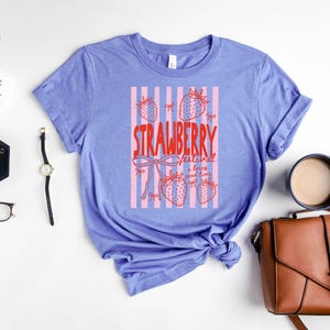 May include: A lilac t-shirt featuring a red graphic that reads "STRAWBERRY festival a berry good time". The design includes pink stripes and strawberry illustrations. The sleeves of the shirt are rolled up.