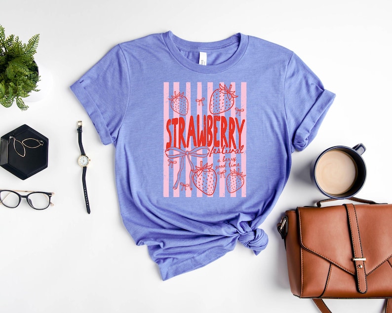 May include: A lilac t-shirt featuring a red graphic that reads "STRAWBERRY festival a berry good time". The design includes pink stripes and strawberry illustrations. The sleeves of the shirt are rolled up.