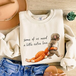 Peut inclure: Sweat-shirt cr&egrave;me avec le texte "Some of us need a little extra love." Le motif repr&eacute;sente un singe brun &eacute;treignant un animal en peluche orange. Le sweat-shirt est associ&eacute; &agrave; un jean bleu, un chapeau beige et des sandales.