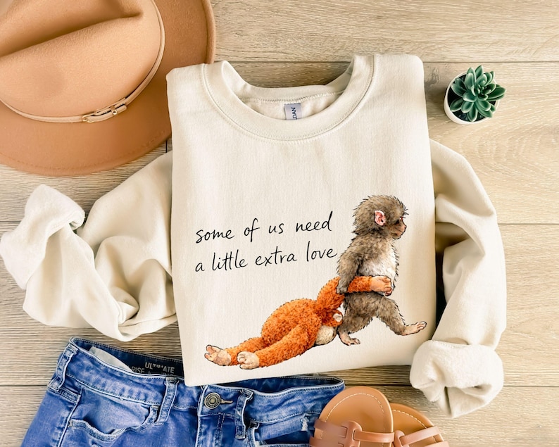Peut inclure: Sweat-shirt cr&egrave;me avec le texte "Some of us need a little extra love." Le motif repr&eacute;sente un singe brun &eacute;treignant un animal en peluche orange. Le sweat-shirt est associ&eacute; &agrave; un jean bleu, un chapeau beige et des sandales.