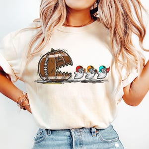 Puede incluir: Camiseta color crema con una calabaza con forma de balón de fútbol con un mordisco, y tres fantasmas de dibujos animados con cascos de fútbol. Los fantasmas sostienen balones de fútbol y llevan cascos rojos, amarillos y azules.