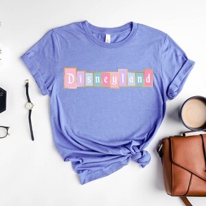 Puede incluir: Una camiseta de color lila claro con la palabra "Disneyland" en un dise&ntilde;o colorido de letras en bloque en el pecho. La camiseta est&aacute; doblada en la parte inferior y rodeada de accesorios, incluyendo un reloj, gafas y un bolso.