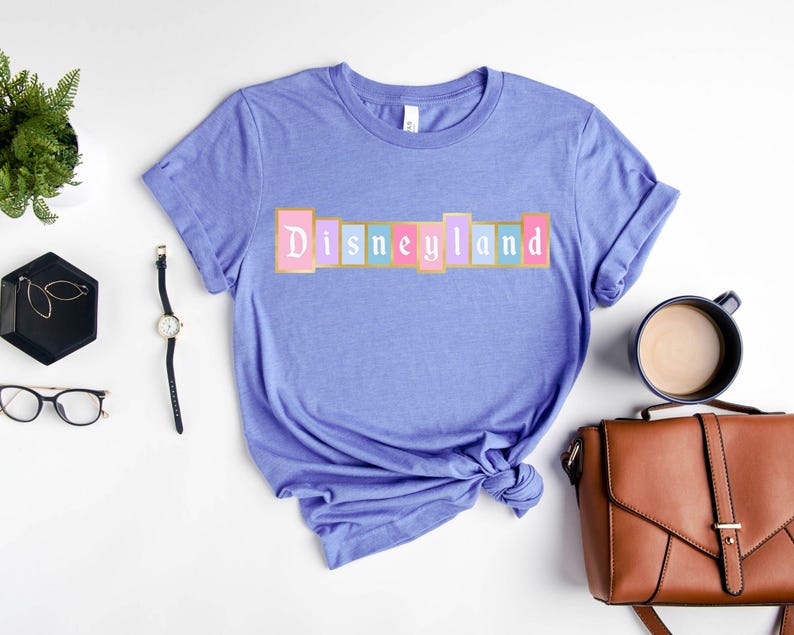 Puede incluir: Una camiseta de color lila claro con la palabra "Disneyland" en un dise&ntilde;o colorido de letras en bloque en el pecho. La camiseta est&aacute; doblada en la parte inferior y rodeada de accesorios, incluyendo un reloj, gafas y un bolso.
