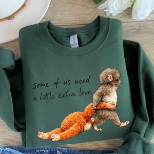 Peut inclure: Un sweat-shirt vert fonc&eacute; avec le texte "some of us need a little extra love". Le sweat-shirt pr&eacute;sente un dessin d'un singe portant un orang-outan. Les manches sont retrouss&eacute;es.