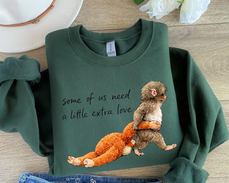 Peut inclure: Un sweat-shirt vert fonc&eacute; avec le texte "some of us need a little extra love". Le sweat-shirt pr&eacute;sente un dessin d'un singe portant un orang-outan. Les manches sont retrouss&eacute;es.