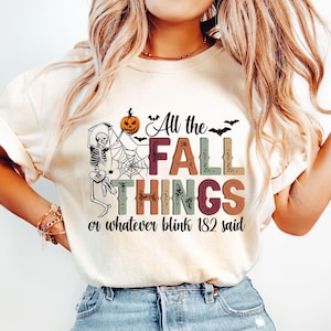 以下が含まれることがあります： ハロウィーンをテーマにしたグラフィックがプリントされたクリーム色のTシャツ。デザインは、骸骨、クモの巣、カボチャ、そして様々な色の「All the FALL THINGS or whatever blink 182 said」というテキストが特徴です。
