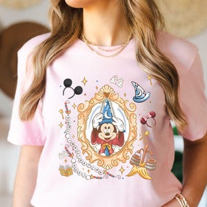 Könnte beinhalten: Hellrosa T-Shirt mit einer gerahmten Illustration von Mickey Mouse mit Zauberhut. Das Design enthält verschiedene Disney-Elemente wie Micky-Maus-Ohren, einen Zauberhut und einen Zauberstab. Das T-Shirt hat kurze Ärmel.