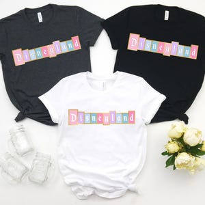 Puede incluir: Tres camisetas en gris oscuro, negro y blanco, cada una con la palabra "Disneyland" en un dise&ntilde;o rectangular colorido. El texto es rosa, azul y amarillo, con contorno dorado. Las camisetas son de manga corta y de estilo casual.