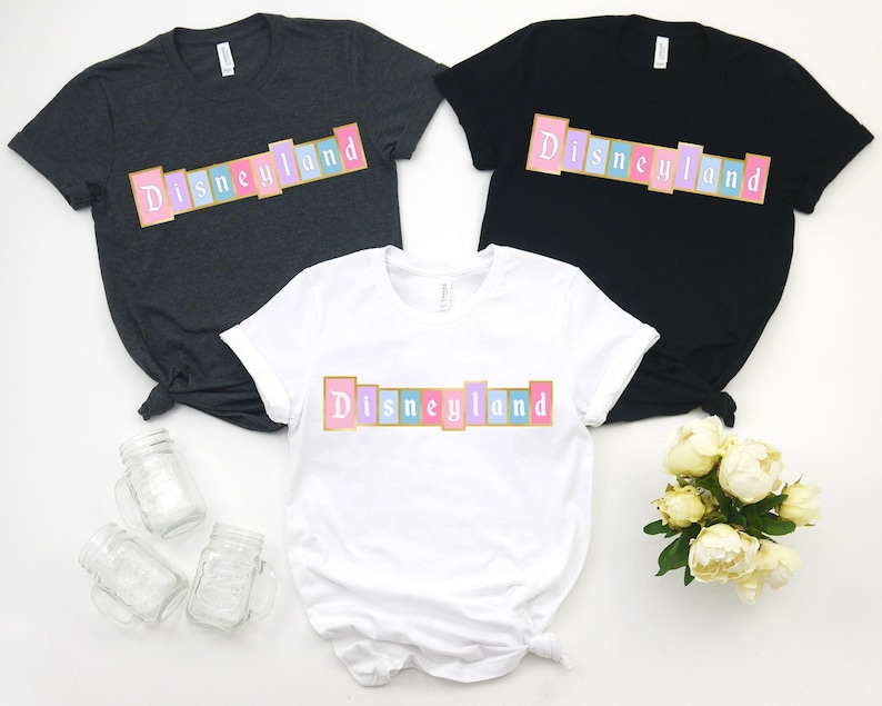Puede incluir: Tres camisetas en gris oscuro, negro y blanco, cada una con la palabra "Disneyland" en un dise&ntilde;o rectangular colorido. El texto es rosa, azul y amarillo, con contorno dorado. Las camisetas son de manga corta y de estilo casual.