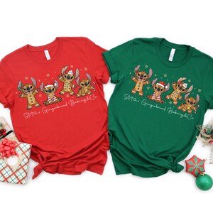 Puede incluir: Dos camisetas de manga corta, una roja y otra verde, con un dise&ntilde;o de dibujos animados de personajes de Stitch como hombres de jengibre. El texto "Stitch's Gingerbread Baking & Co" est&aacute; impreso debajo. Se ven decoraciones navide&ntilde;as.