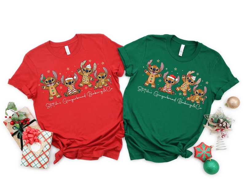 Puede incluir: Dos camisetas de manga corta, una roja y otra verde, con un dise&ntilde;o de dibujos animados de personajes de Stitch como hombres de jengibre. El texto "Stitch's Gingerbread Baking & Co" est&aacute; impreso debajo. Se ven decoraciones navide&ntilde;as.