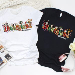 May include: Two t-shirts, one white and one black, with a colorful Disneyland Christmas graphic. The graphic features Disney characters and the words "Disneyland Christmas" in a festive font. The shirts are laid flat on a white surface.