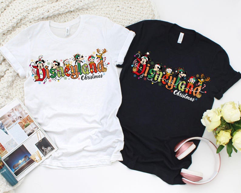 May include: Two t-shirts, one white and one black, with a colorful Disneyland Christmas graphic. The graphic features Disney characters and the words "Disneyland Christmas" in a festive font. The shirts are laid flat on a white surface.