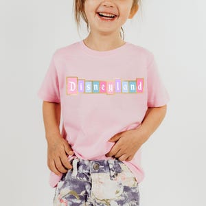Puede incluir: Una camiseta rosa claro con la palabra "Disneyland" impresa en el pecho en un dise&ntilde;o colorido de letras en bloque. Las letras est&aacute;n delineadas en dorado, con bloques rosas, azules y amarillos. La camiseta la lleva un ni&ntilde;o.