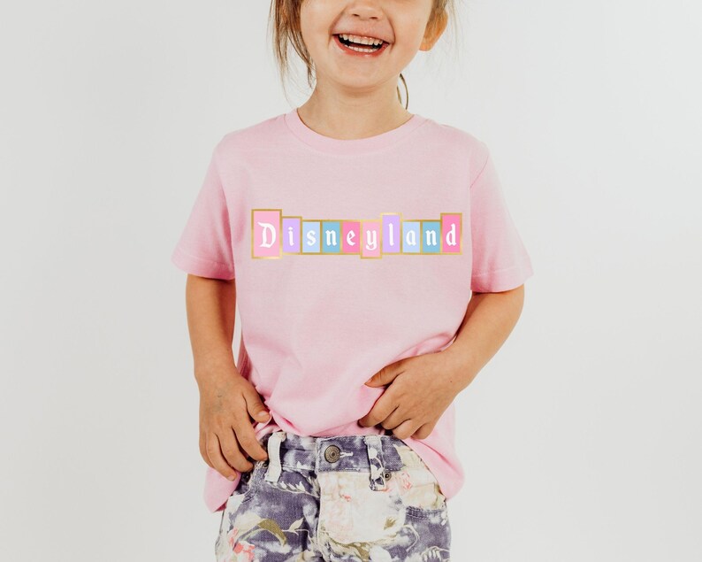 Puede incluir: Una camiseta rosa claro con la palabra "Disneyland" impresa en el pecho en un dise&ntilde;o colorido de letras en bloque. Las letras est&aacute;n delineadas en dorado, con bloques rosas, azules y amarillos. La camiseta la lleva un ni&ntilde;o.
