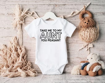 May include: A white baby onesie with the text "TAKE ME TO MY AUNT YOU PEASANT" in black. The onesie is hanging on a white hanger. The background includes decorative items like a teddy bear and a woven ball.