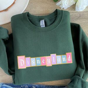 Puede incluir: Una sudadera verde oscuro con la palabra "Disneyland" en una fuente retro colorida. El texto est&aacute; sobre una serie de bloques rectangulares en rosa, morado y azul. La sudadera est&aacute; doblada, con las mangas visibles.