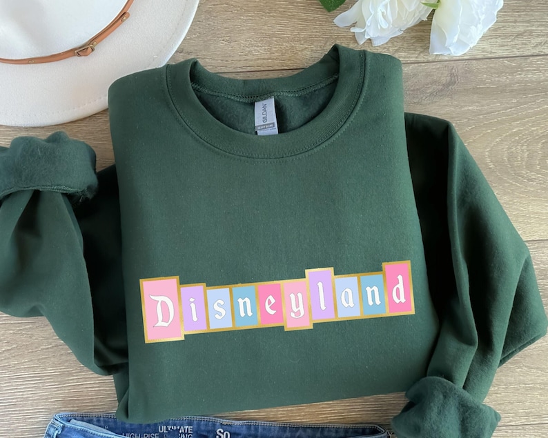 Puede incluir: Una sudadera verde oscuro con la palabra "Disneyland" en una fuente retro colorida. El texto est&aacute; sobre una serie de bloques rectangulares en rosa, morado y azul. La sudadera est&aacute; doblada, con las mangas visibles.