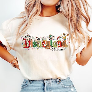 May include: Cream-colored t-shirt featuring the text "Disneyland Christmas" in a colorful, festive design. The design includes Mickey Mouse, Donald Duck, Goofy, and other characters in holiday attire. The shirt is a casual, comfortable style.