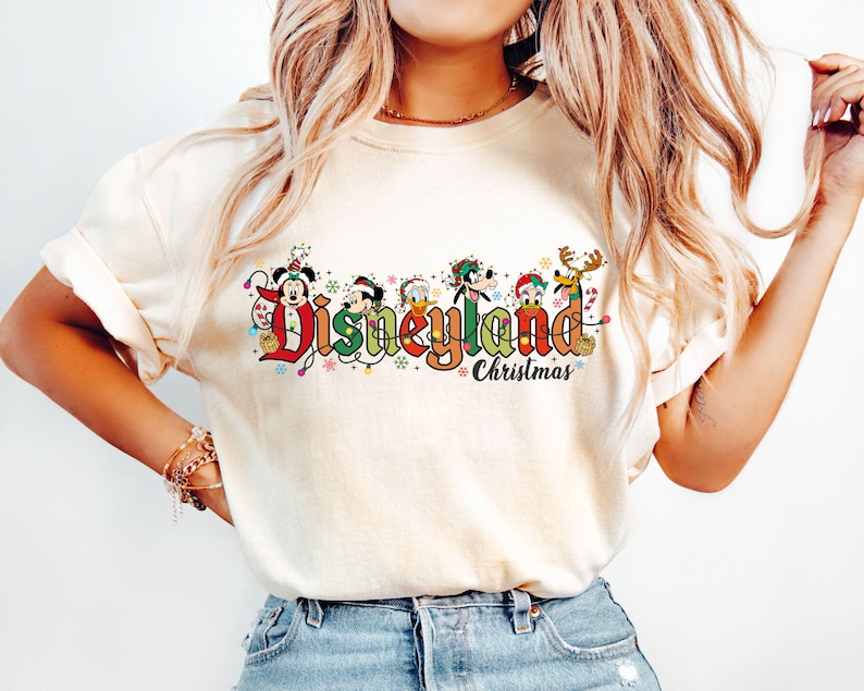 May include: Cream-colored t-shirt featuring the text "Disneyland Christmas" in a colorful, festive design. The design includes Mickey Mouse, Donald Duck, Goofy, and other characters in holiday attire. The shirt is a casual, comfortable style.