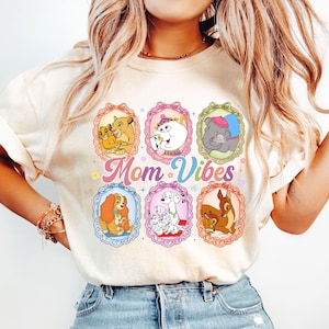 Può includere: T-shirt color crema con sei immagini di cartoni animati di personaggi Disney incorniciate. Le parole "Mom Vibes" sono stampate in caratteri colorati. La maglietta è realizzata in materiale morbido.