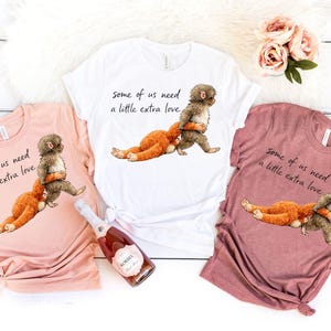 Peut inclure: Trois t-shirts roses, blancs et mauves, chacun avec une illustration fantaisiste d'une cr&eacute;ature en train d'embrasser une autre. Le texte "Some of us need a little extra love" est imprim&eacute; sur chaque chemise. Une bouteille de champagne Korbel est &eacute;galement visible.