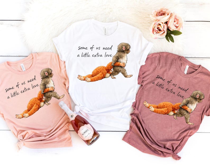 Peut inclure: Trois t-shirts roses, blancs et mauves, chacun avec une illustration fantaisiste d'une cr&eacute;ature en train d'embrasser une autre. Le texte "Some of us need a little extra love" est imprim&eacute; sur chaque chemise. Une bouteille de champagne Korbel est &eacute;galement visible.