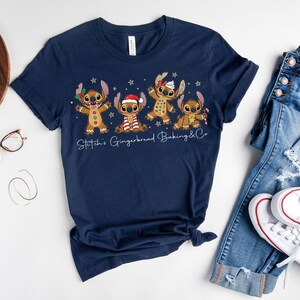 Puede incluir: Camiseta azul marino con un dise&ntilde;o festivo de cuatro personajes de jengibre con accesorios navide&ntilde;os. El texto "Stitch's Gingerbread Baking & Co" est&aacute; impreso debajo. La camiseta se muestra con vaqueros azules y zapatillas blancas.