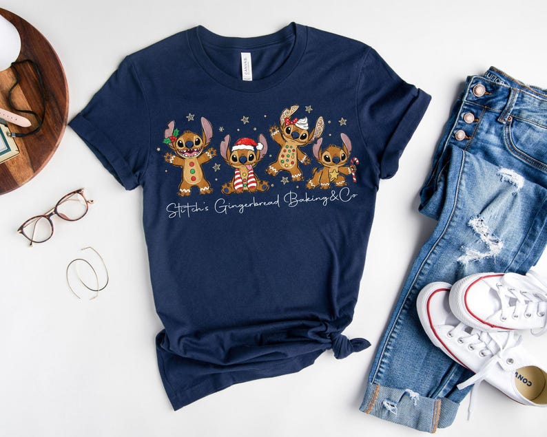 Puede incluir: Camiseta azul marino con un dise&ntilde;o festivo de cuatro personajes de jengibre con accesorios navide&ntilde;os. El texto "Stitch's Gingerbread Baking & Co" est&aacute; impreso debajo. La camiseta se muestra con vaqueros azules y zapatillas blancas.